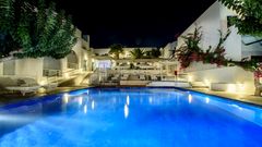 Stimmungsvoll beleuchteter Hotelpool bei Nacht – weiße Gebäude im Kykladenstil, umgeben von Palmen, Bougainvillea und gemütlichen Liegen.
