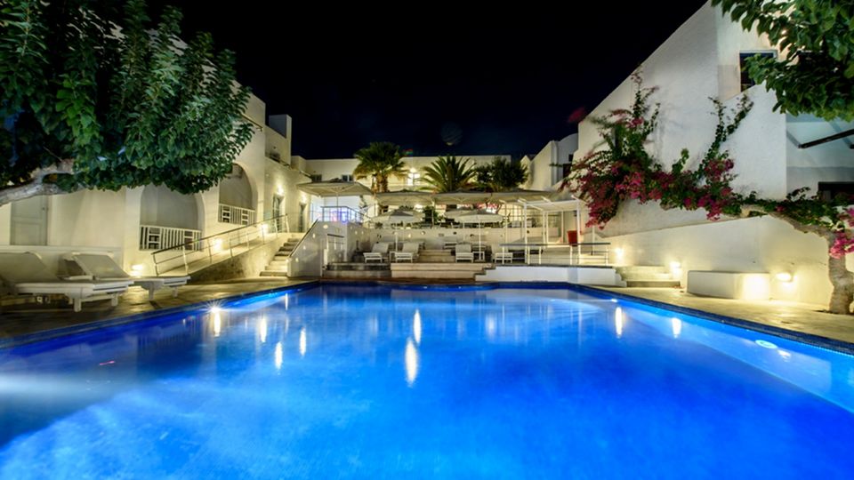 Stimmungsvoll beleuchteter Hotelpool bei Nacht – weiße Gebäude im Kykladenstil, umgeben von Palmen, Bougainvillea und gemütlichen Liegen.