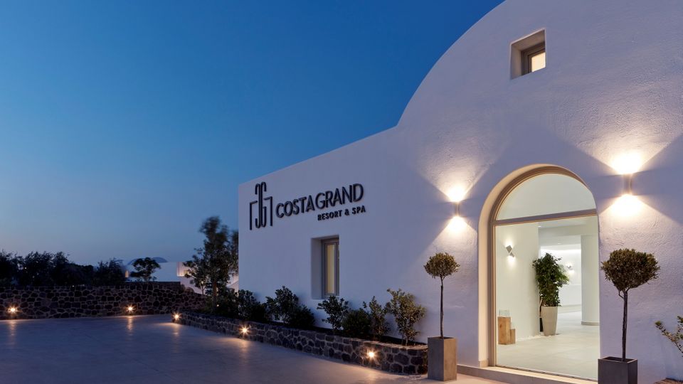 Beleuchteter Hoteleingang des Costa Grand Resort & Spa am Abend – modernes, weißes Gebäude im Kykladenstil mit stilvoller Außenbeleuchtung und mediterraner Bepflanzung.