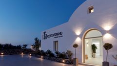 Beleuchteter Hoteleingang des Costa Grand Resort & Spa am Abend – modernes, weißes Gebäude im Kykladenstil mit stilvoller Außenbeleuchtung und mediterraner Bepflanzung.